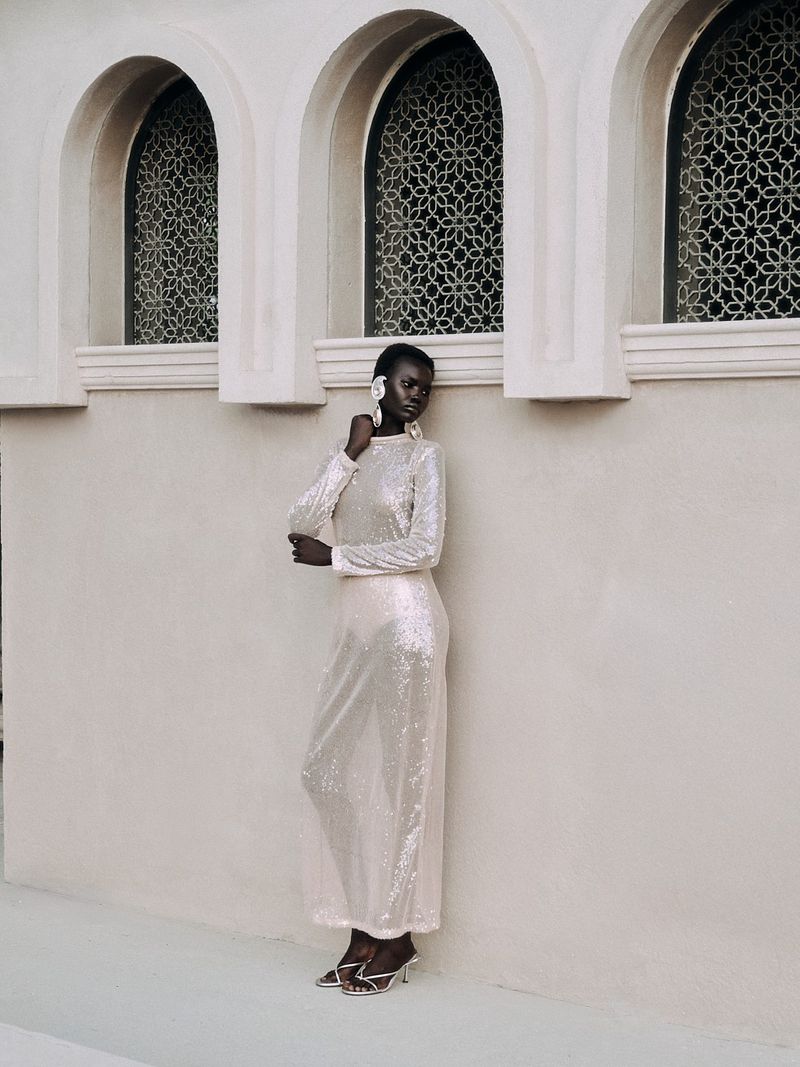 An African woman in a full-length shimmering gown poses near arched windows in an outdoor setting, showcasing fashion and beauty.