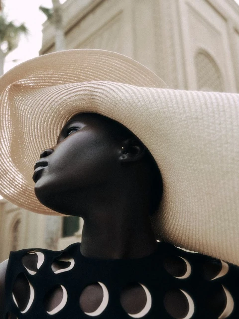 Elegant dark-skinned woman in black dress with moon cutouts, wearing a large sun hat against a light-colored, classical building backdrop.