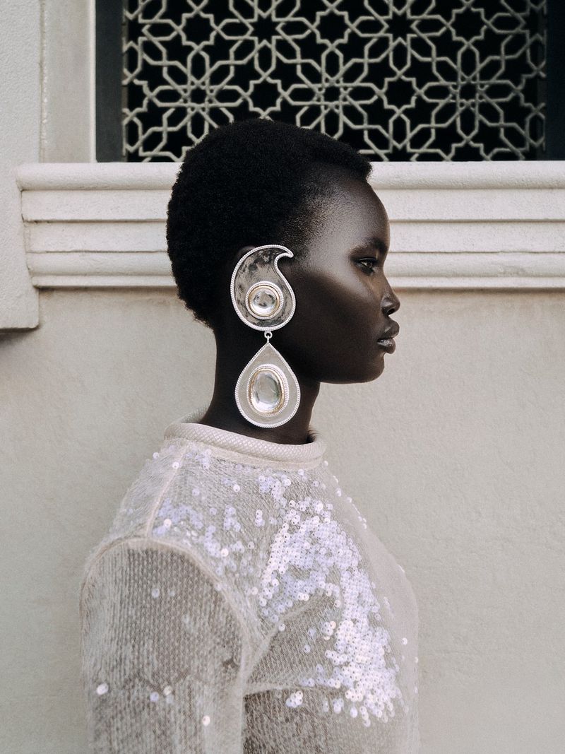 Profile shot of a Black woman wearing oversized silver teardrop earrings and a sparkling light-colored sweater against a neutral background.
