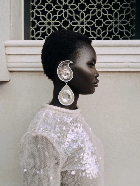 Profile shot of a Black woman wearing oversized silver teardrop earrings and a sparkling light-colored sweater against a neutral background.