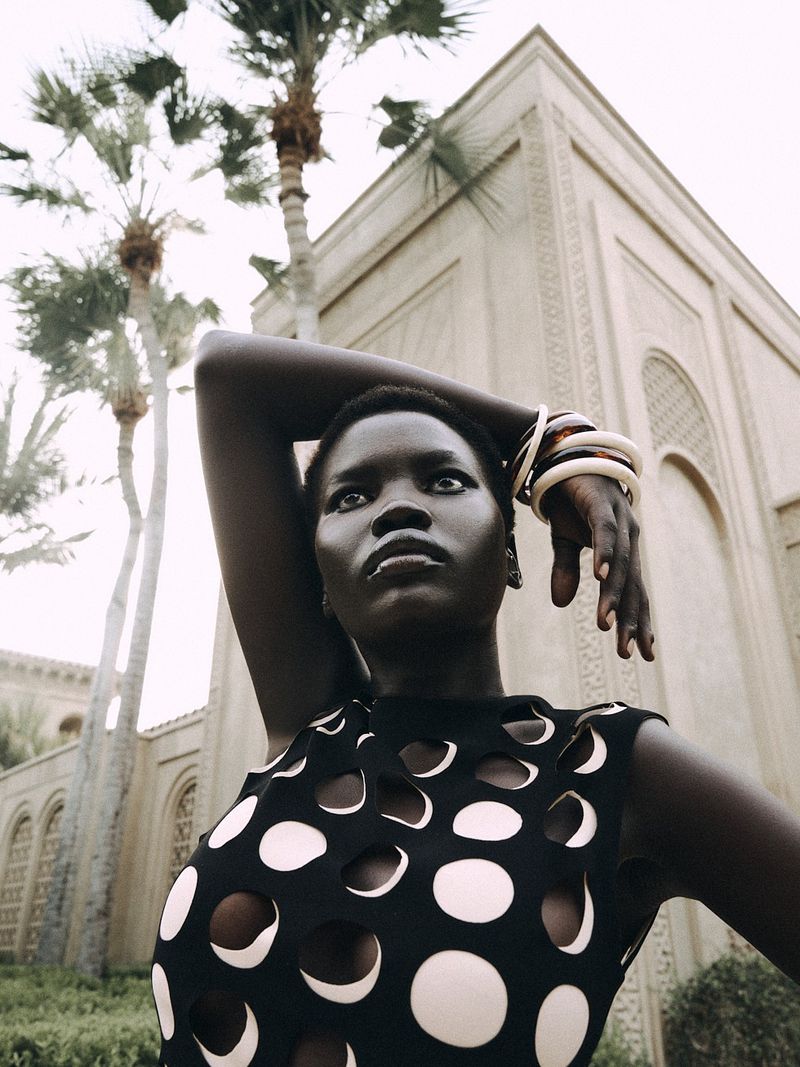 Fashion shot of a Black woman with jewelry posing in a sleeveless top with circle cutouts, set against a building and palm trees.