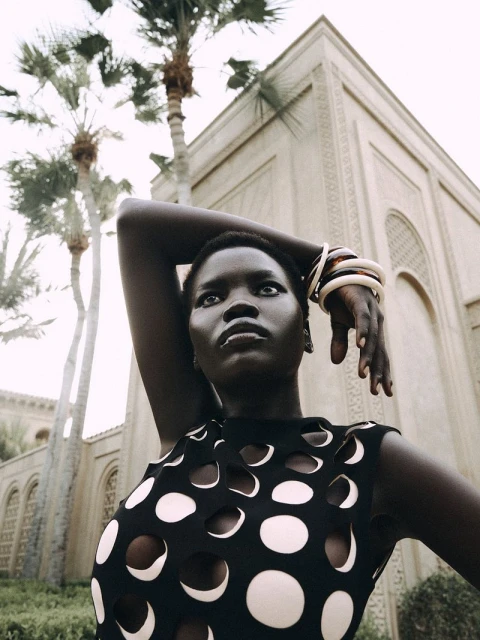 Fashion shot of a Black woman with jewelry posing in a sleeveless top with circle cutouts, set against a building and palm trees.