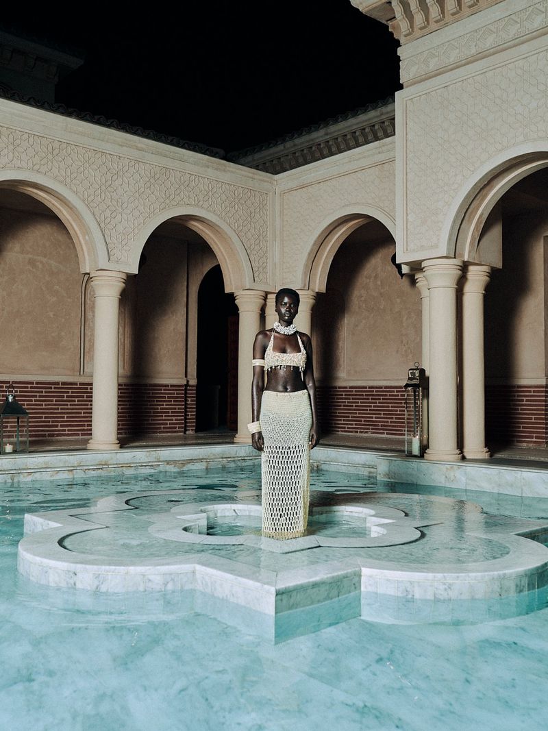 A tall, dark-skinned woman stands in a light-blue tile pool with beige arches and columns. She wears a crocheted, semi-sheer outfit.