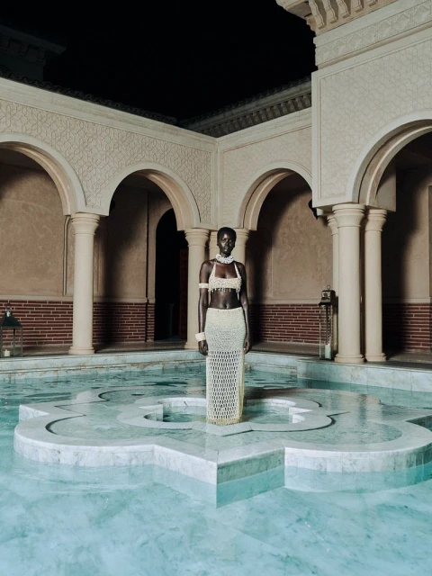 A tall, dark-skinned woman stands in a light-blue tile pool with beige arches and columns. She wears a crocheted, semi-sheer outfit.