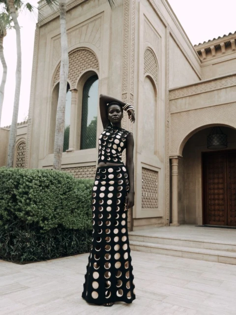 A dark-skinned woman in patterned black-and-beige dress poses in front of a light-stone building with arched openings and green shrubbery.