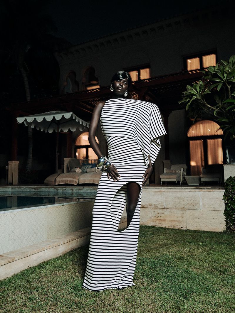 A stylish dark-skinned woman models a striped, asymmetrical dress outside a mansion with a pool, at night. The setting is luxurious and refined.
