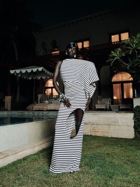 A stylish dark-skinned woman models a striped, asymmetrical dress outside a mansion with a pool, at night. The setting is luxurious and refined.