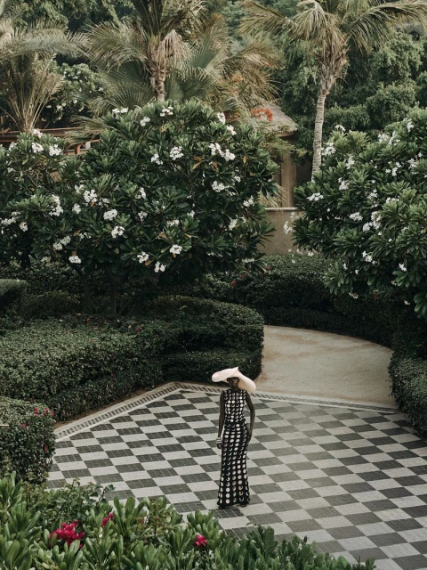 Woman in patterned dress and large hat stands on checkered tile in a lush tropical garden, surrounded by green foliage and palm trees.