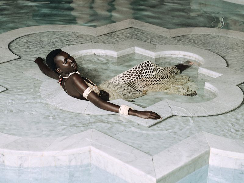 Adult woman lies in shallow water of a marble pool wearing a crocheted dress and jewelry, in a medium shot, indoor, in ambient lighting.