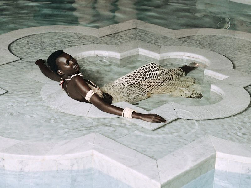 Adult woman lies in shallow water of a marble pool wearing a crocheted dress and jewelry, in a medium shot, indoor, in ambient lighting.