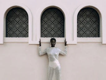 Black woman wearing a sheer silver sequin dress posing against a pale wall with three arched windows for oothepalm x afrobougee