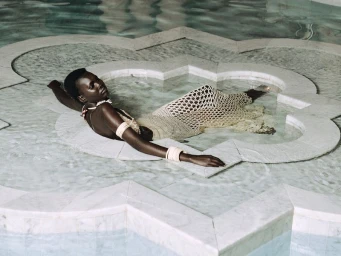 Black woman lounging in an ornate geometric marble pool wearing a netted crochet dress and bracelets.