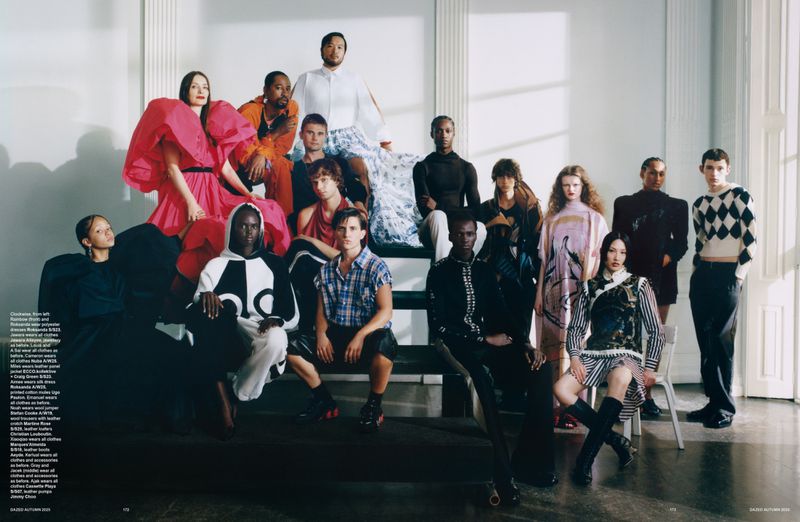 Fashion editorial shot featuring a large group of diverse models in various outfits, posing on stairs and platforms in a bright indoor setting.