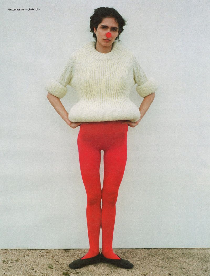 An adult woman poses in a studio wearing an oversized sweater, red tights, ballet flats, and a red clown nose against a white wall.