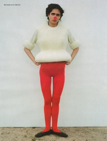 An adult woman poses in a studio wearing an oversized sweater, red tights, ballet flats, and a red clown nose against a white wall.