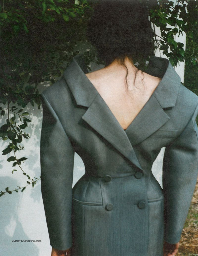 Back view of a woman in a structured gray blazer dress with squared shoulders outdoors against a background of greenery and a painted wall.