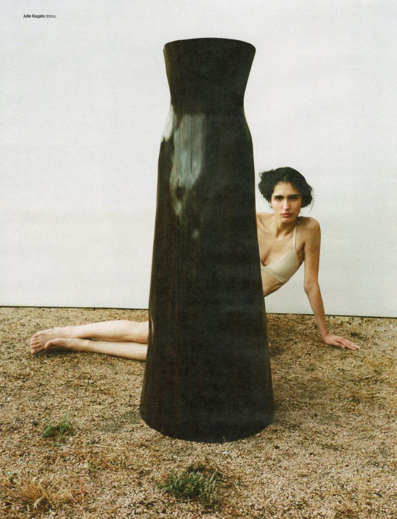 Woman poses beside a large, sculpted black dress on a tan dirt surface against a neutral background in a fashion editorial shot.