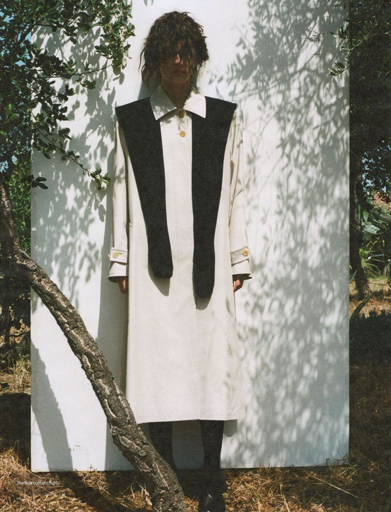 Fashion model posing outdoors wearing a beige coat with a large black shawl-like collar against a white wall backdrop with tree branches.