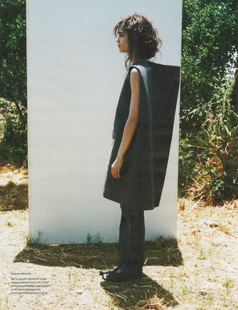 A woman stands in a minimalist sleeveless dress in an outdoor setting against a partial white wall, creating a fashion editorial image.