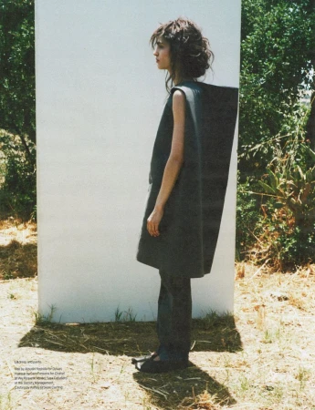 A woman stands in a minimalist sleeveless dress in an outdoor setting against a partial white wall, creating a fashion editorial image.