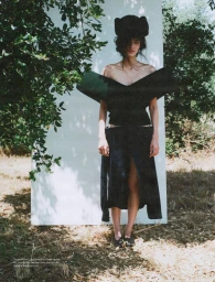 Model wearing a black structured top and skirt with a large black hat standing in front of a white panel outdoors
