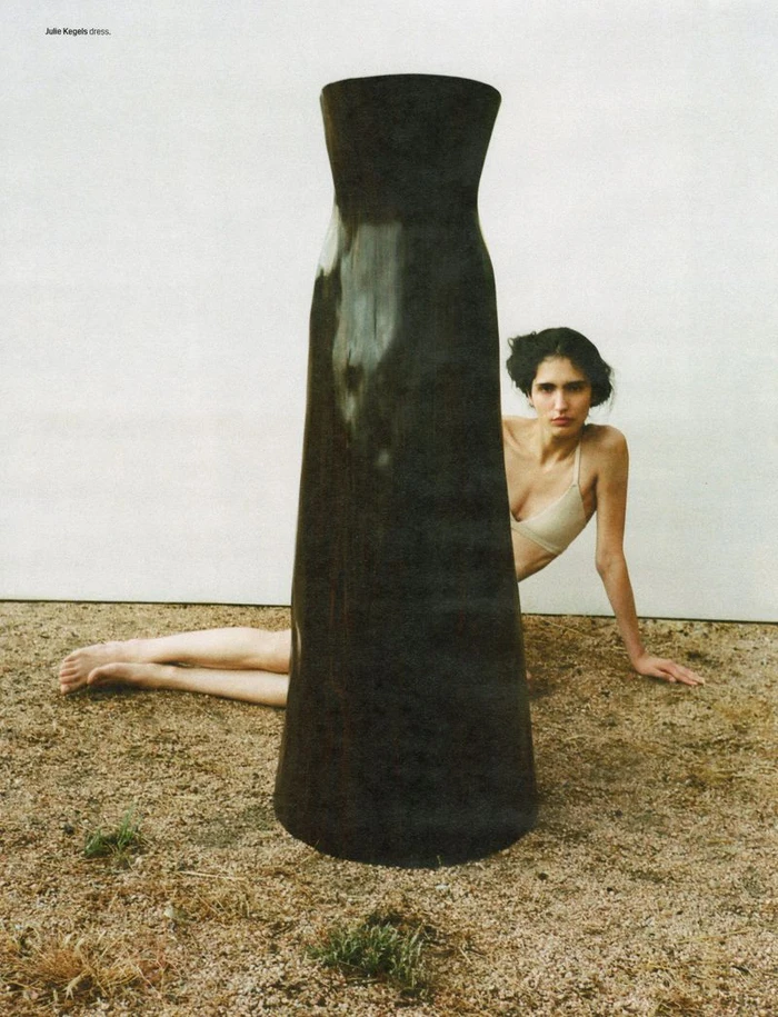 Model seated on the ground behind a large black sculptural column with sand floor and white wall background