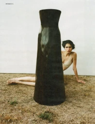 Model seated on the ground behind a large black sculptural column with sand floor and white wall background