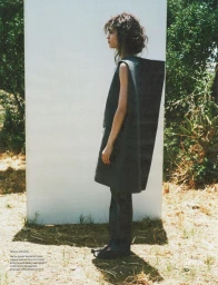 Profile shot of model wearing a sculptural dark tunic standing outdoors beside a white panel against trees.