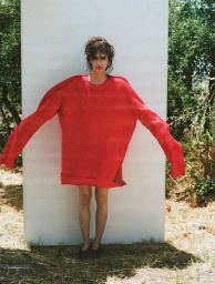 Model wearing an oversized red sweater standing against a white panel outdoors in a green environment.
