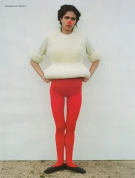 Model wearing a sculptural cream chunky knit sweater, bright red tights, black flats, and a red nose against a plain white background.