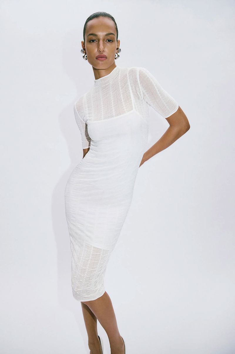 A slender woman poses in a semi-sheer white dress against a bright white backdrop in a fashion or beauty shot with studio lighting.