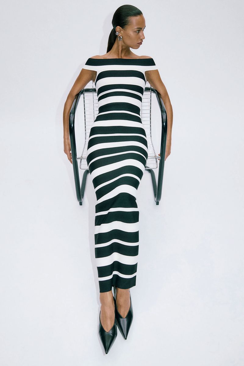 Woman sits in a chair wearing a black-and-white striped dress and black heels against a white background in a fashion studio shot.