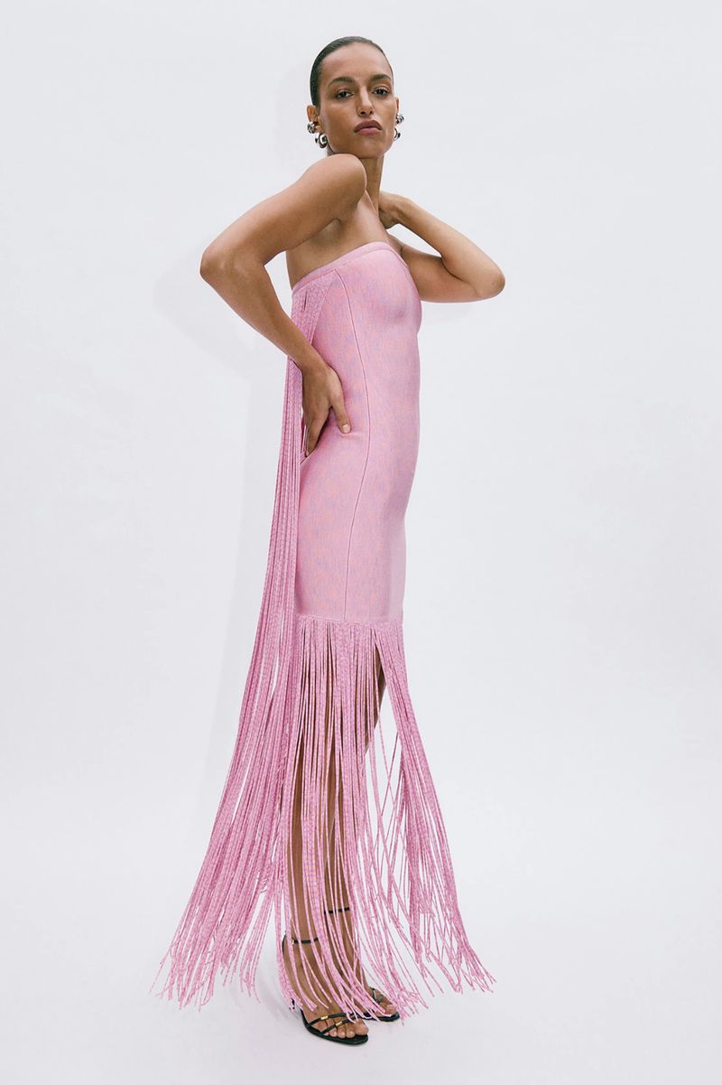 Elegant adult female in a pink strapless dress with fringe and black high heels stands in a photo studio with a neutral white background.