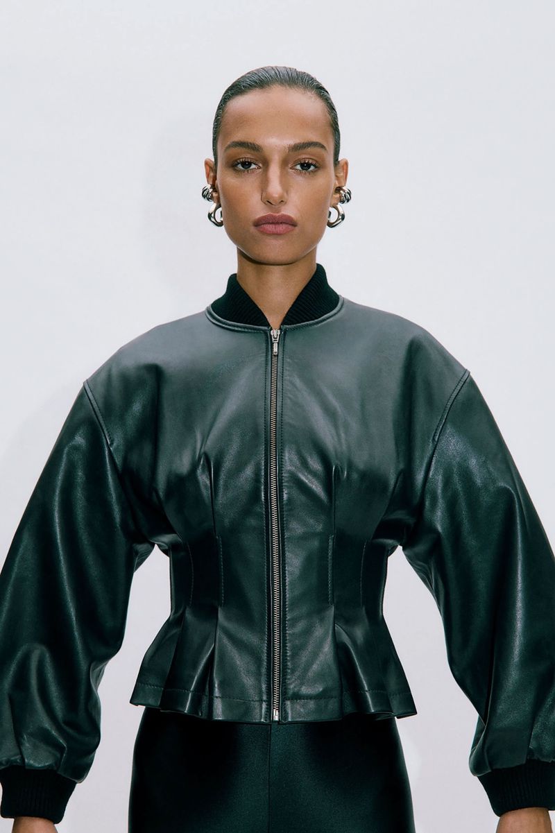 Portrait of a woman wearing a dark green leather bomber jacket with puffed sleeves and chunky silver hoop earrings against white background.
