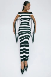 Model wearing a black and white striped off-the-shoulder maxi dress seated in a modern metal chair against a white background.