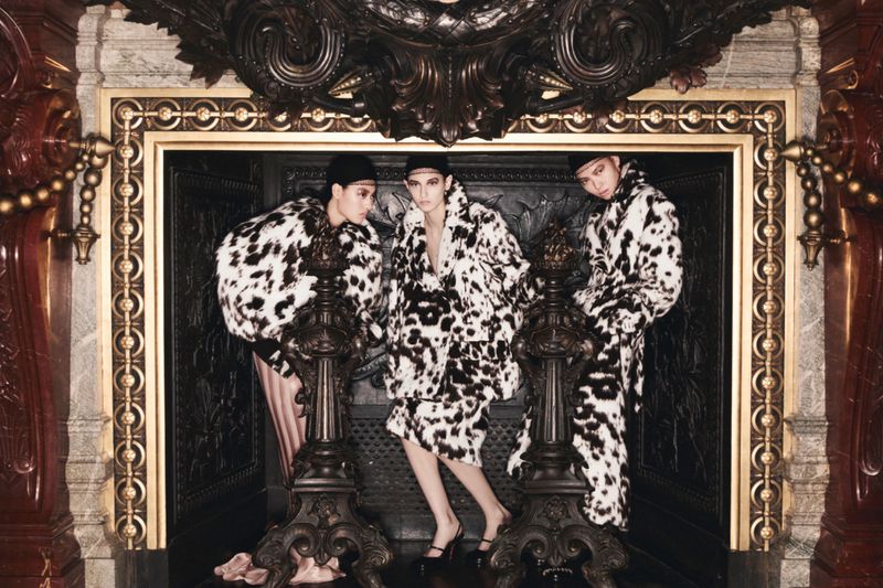 Three fashion models wearing black and white patterned coats pose in front of an ornate, decorative fireplace in a studio setting.