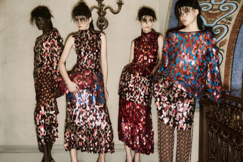 Four fashion models pose indoors wearing sequined dresses and patterned pants. The models stand in front of a tiled wall and chandelier.