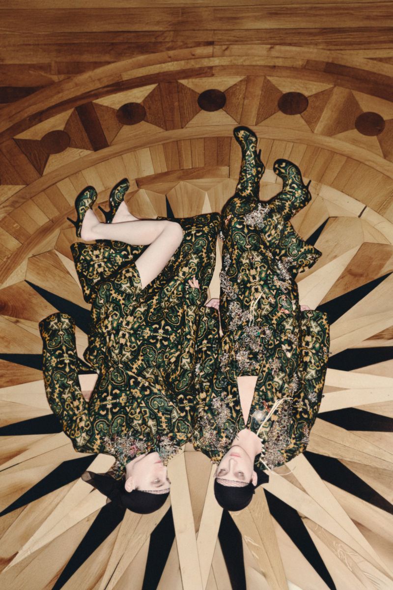 Two fashion models wearing matching patterned outfits and heeled boots are lying head-to-head on a starburst wood floor indoors.
