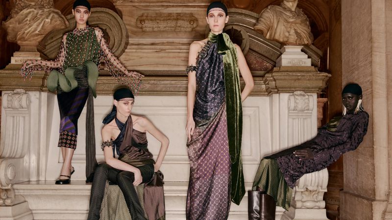 Four fashion models in draped dresses pose against a backdrop of classical architectural details and sculpture in a staged studio shot.