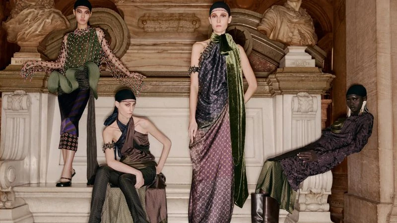 Four fashion models in draped dresses pose against a backdrop of classical architectural details and sculpture in a staged studio shot.