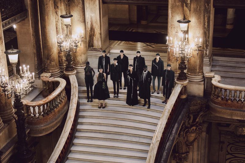 A group of models pose on the stairs of a grand marble staircase in a dimly lit, ornate building with chandeliers. Fashion shoot.