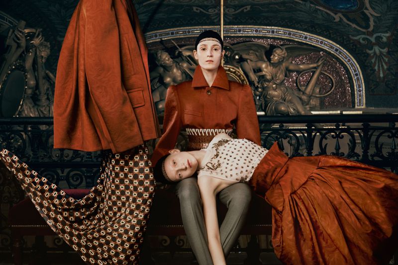 Two women in a fashion portrait with ornate background, one woman sitting with the other leaning across her lap, both in orange tones.