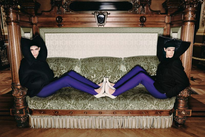 Two women mirrored on a decorative green couch, wearing black hooded tops and purple leggings, indoors on a wood floor with carved wood elements.