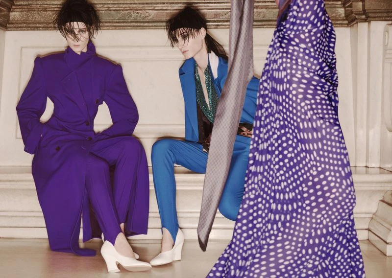 Two models in vibrant purple and blue tailored suits seated against a classical stone interior