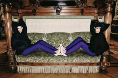 Two models in black hooded jackets and purple tights reclining symmetrically on an ornate green fringed sofa.