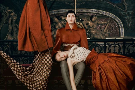 Two models in rust-colored structured garments posed against an ornate classical interior with sculptural reliefs