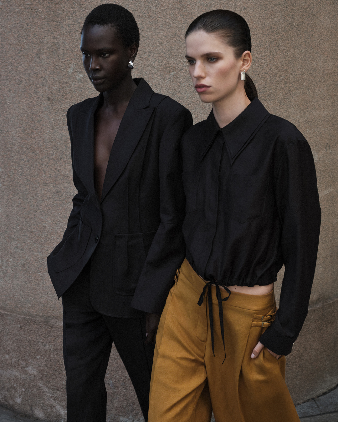 Two fashion models pose in black tops and neutral pants against a textured wall, exuding elegance and style in a studio setting.