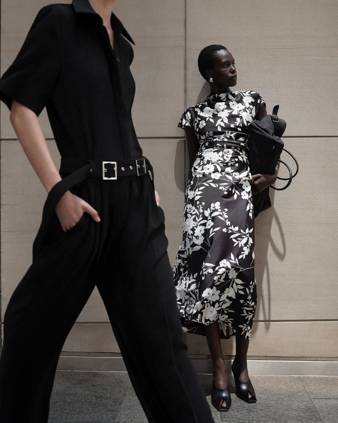 Two models posing outdoors against a stone wall for Cue Autumn 25, wearing a black jumpsuit and a black and white floral dress.