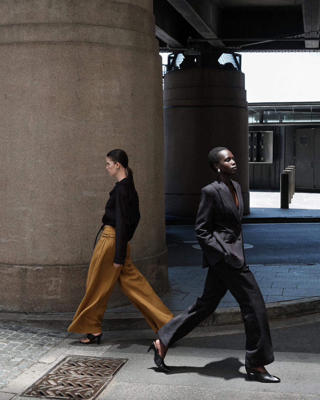 Two women stride confidently in a fashion editorial. One in yellow pants and the other in a dark suit under an elevated structure.