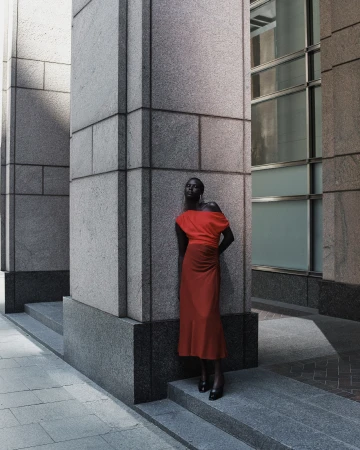 An African-American woman in a red dress poses against stone columns in an urban setting, with architectural details in the background.